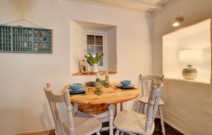 A dining area with a wooden table, blue bowls, white chairs, a vase with flowers, and a wall-mounted shelf with decorative items.