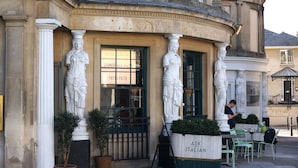 A building with classical architecture, featuring columns and statues. There are outdoor seating areas with tables and chairs.