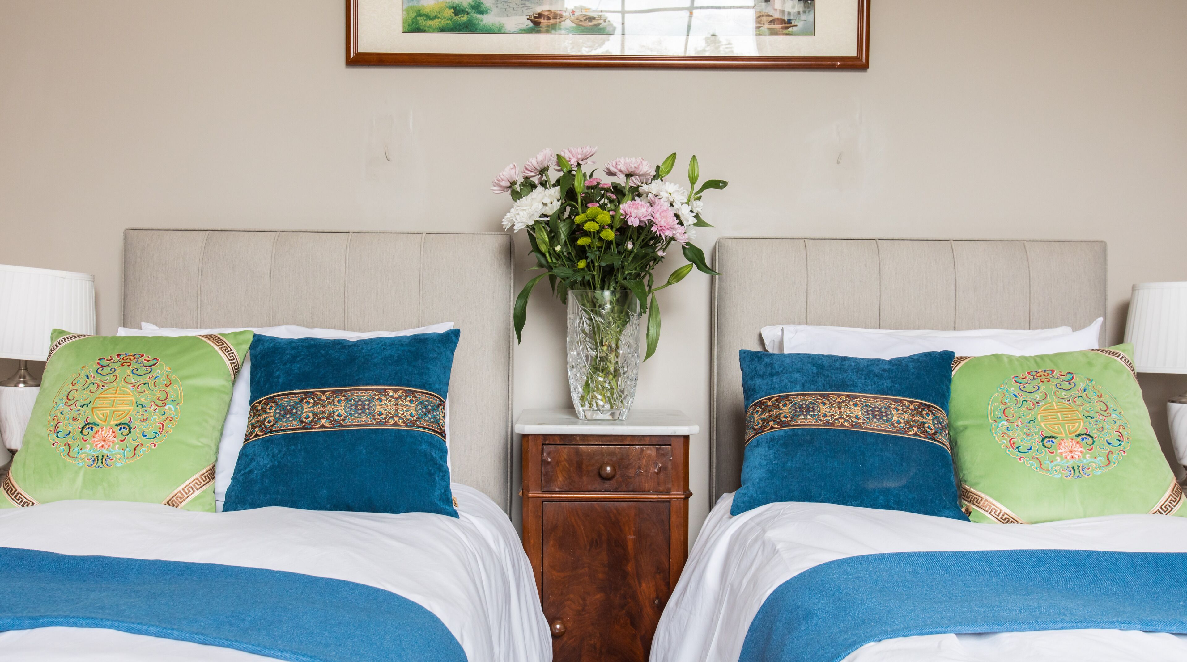 Ein Hotelzimmer mit zwei Betten, jedes mit blaugrünen Zierkissen, einem Holfnachtkästchen mit Blumen in einer Vase und einem gerahmten Bild an der Wand.