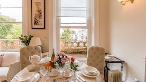 A dining room with a table set for four, featuring white plates, glasses, and a bottle of wine. There are two chairs, a vase with flowers, and a lamp on the table.