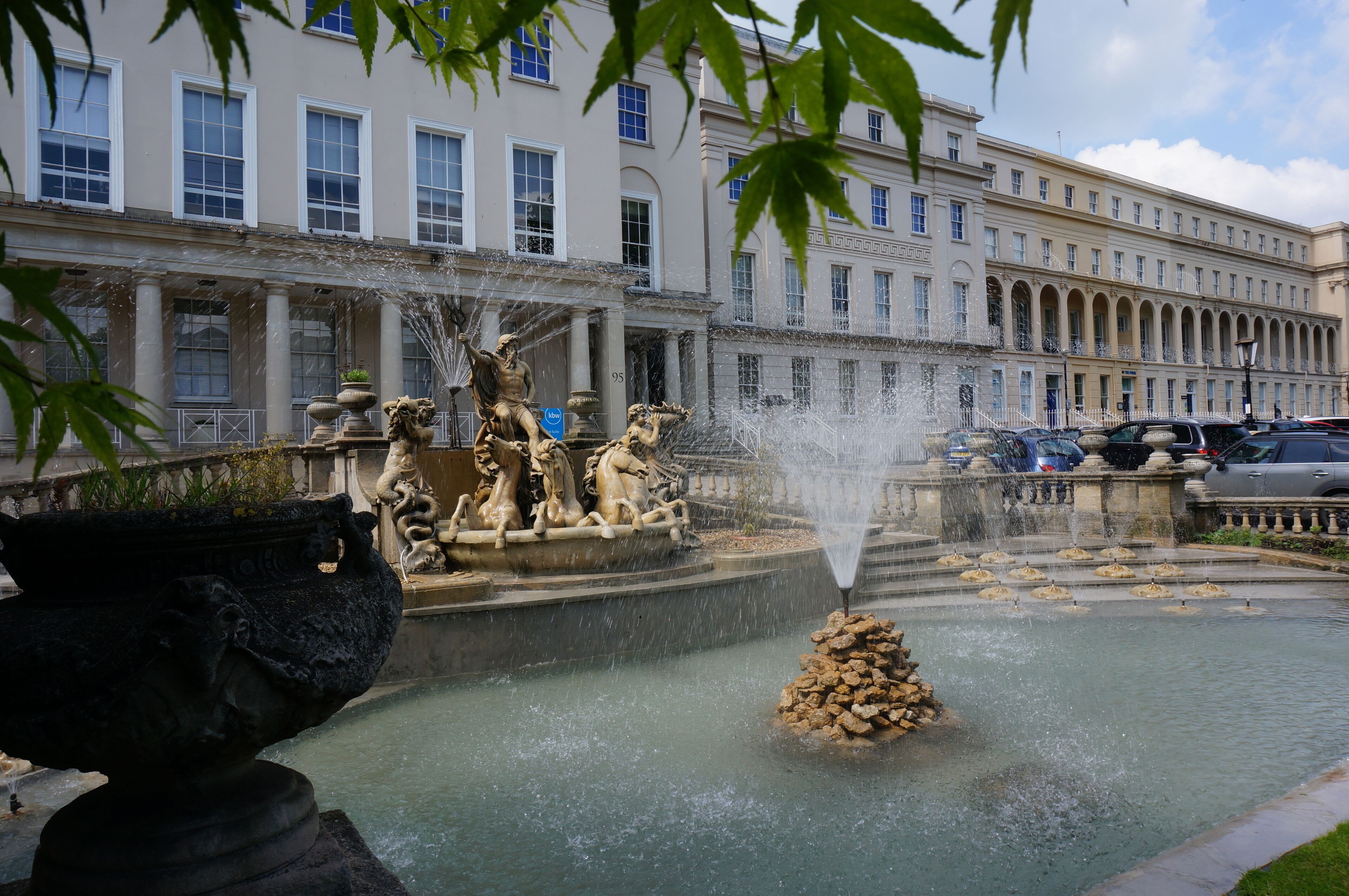 Une fontaine classique, ornée de statues et de jets d’eau, se trouve devant un grand bâtiment doté de nombreuses fenêtres et de colonnes.