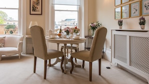 A dining area with a round table set for four, surrounded by four chairs. There is a radiator on the right side and a window with a view of buildings outside.