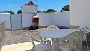 Um terraço no telhado com uma mesa, cadeiras, churrasqueira e uma parede branca com uma porta.