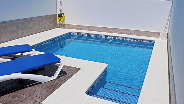 Uma piscina pequena e retangular, com borda azulejada, uma mangueira e duas cadeiras de descanso azuis.