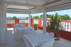 An outdoor patio with white tables and chairs, potted plants, and a view of the sea. - Hostal Amanecer (Cienfuegos)