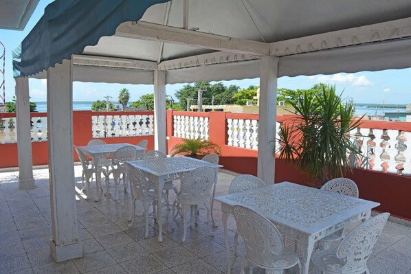 An outdoor patio with white tables and chairs, a red railing, and a view of the sea. - Hostal Amanecer (Cienfuegos)