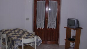 A room with a checkered table, a wooden chair, a TV on a wooden stand, and a door with lace curtains.