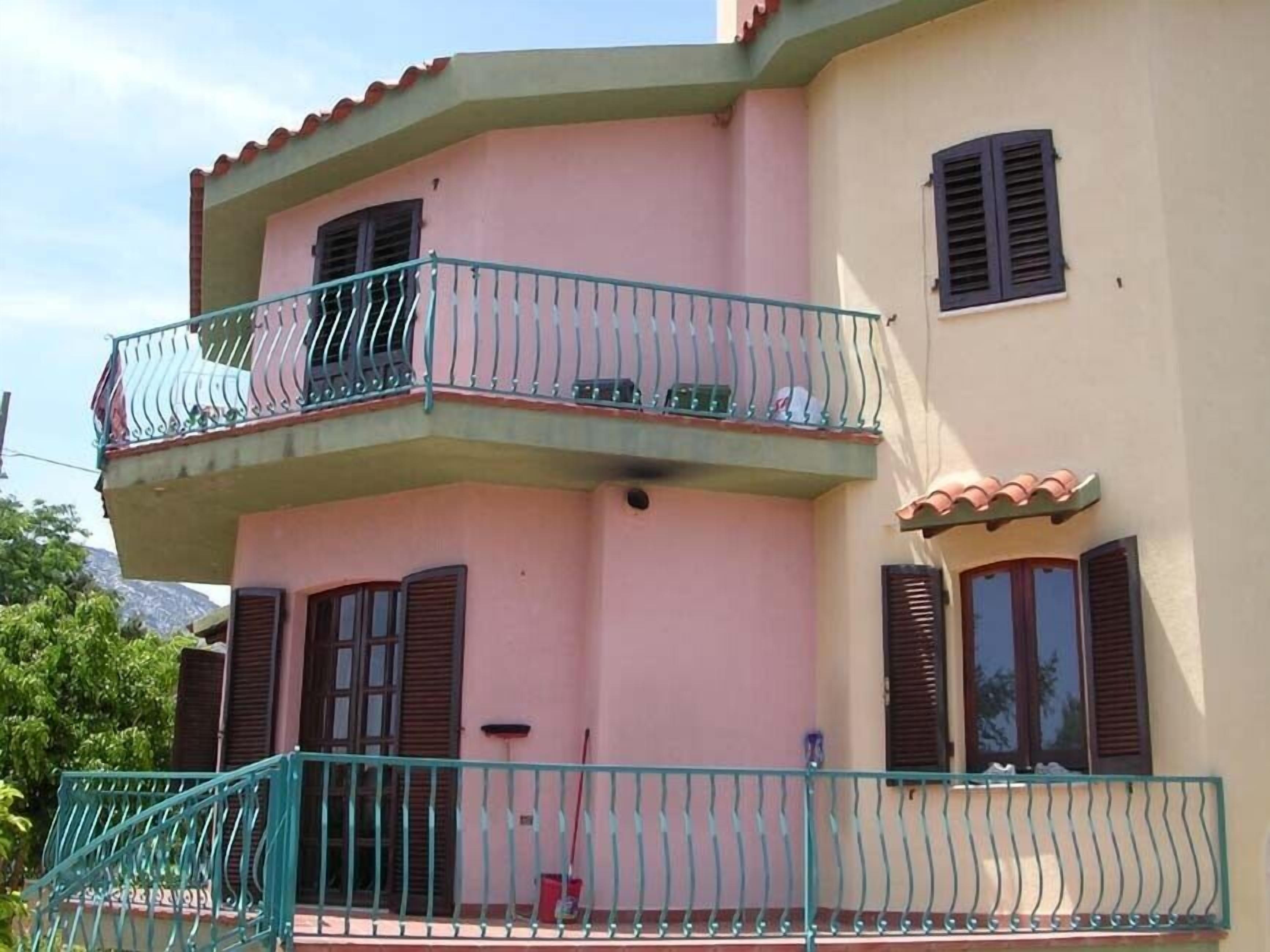 Un edificio a due piani con facciata rosa, un balcone con ringhiere in metallo e persiane alle finestre.
