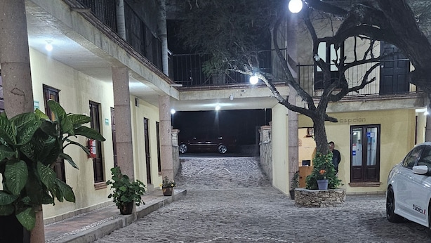 A stone-paved courtyard with a car parked on the right, potted plants, and a building with a balcony in the background. - Hotel Villa Aurora (Ezequiel Montes)