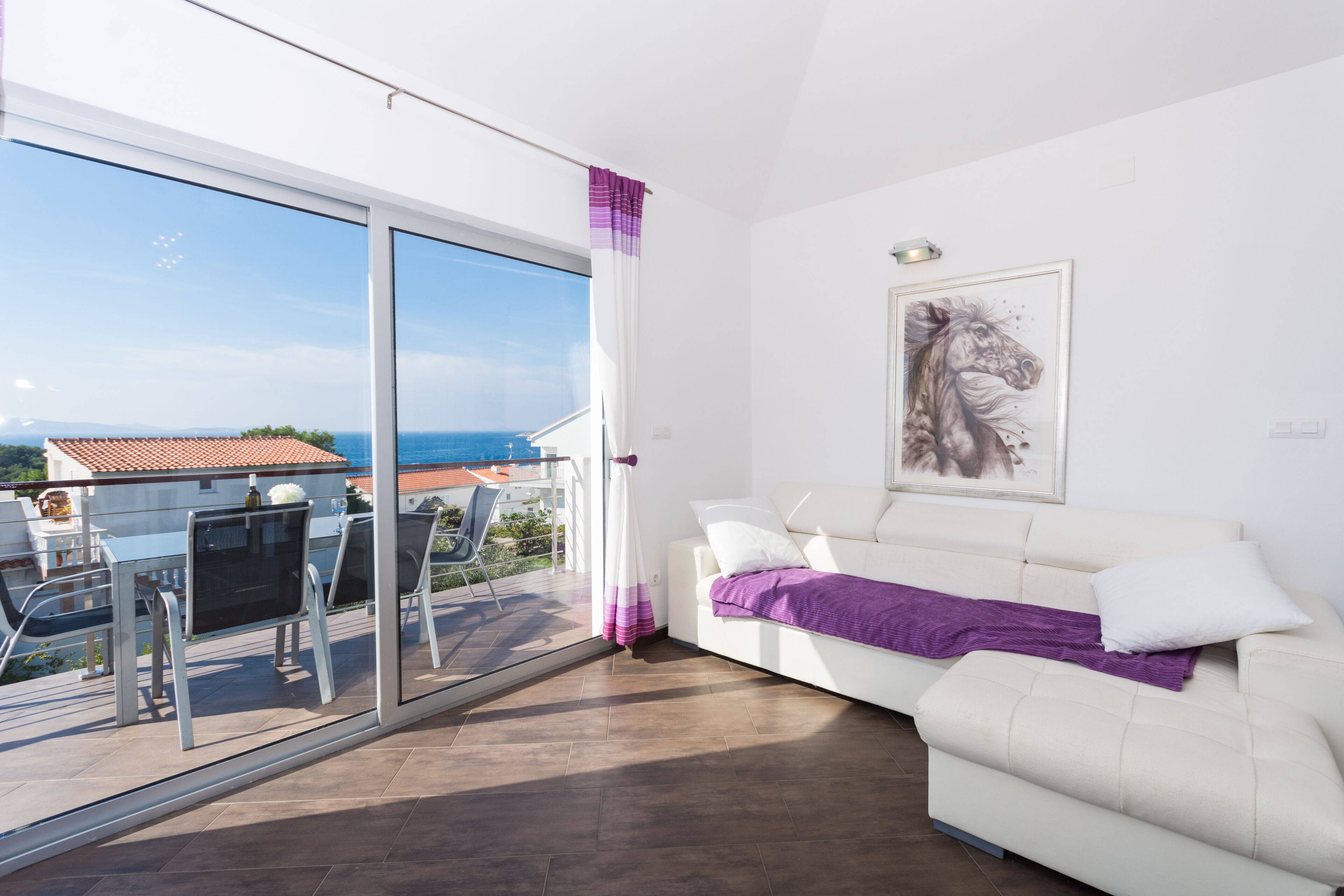 Een moderne woonkamer met een witte bank, een glazen deur naar een balkon en een lijstwerk kunstwerk aan de muur.