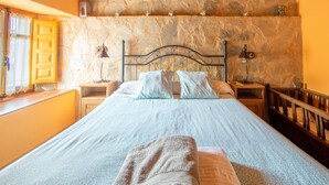 A bedroom with a stone wall, a bed with two pillows, a nightstand with a lamp, and a window with curtains. - Tranquil Stay Near Luna River (Benllera, Carrocera, León)