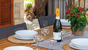 A wooden table set for dining with white plates, glasses, and a bottle of wine, surrounded by wicker baskets and greenery.