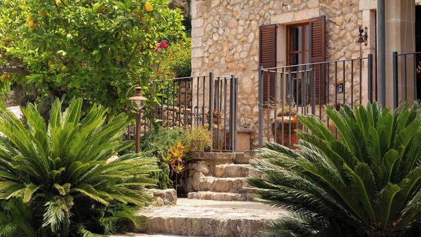 A stone building with a staircase leading to a wooden door, surrounded by lush greenery and palm trees.