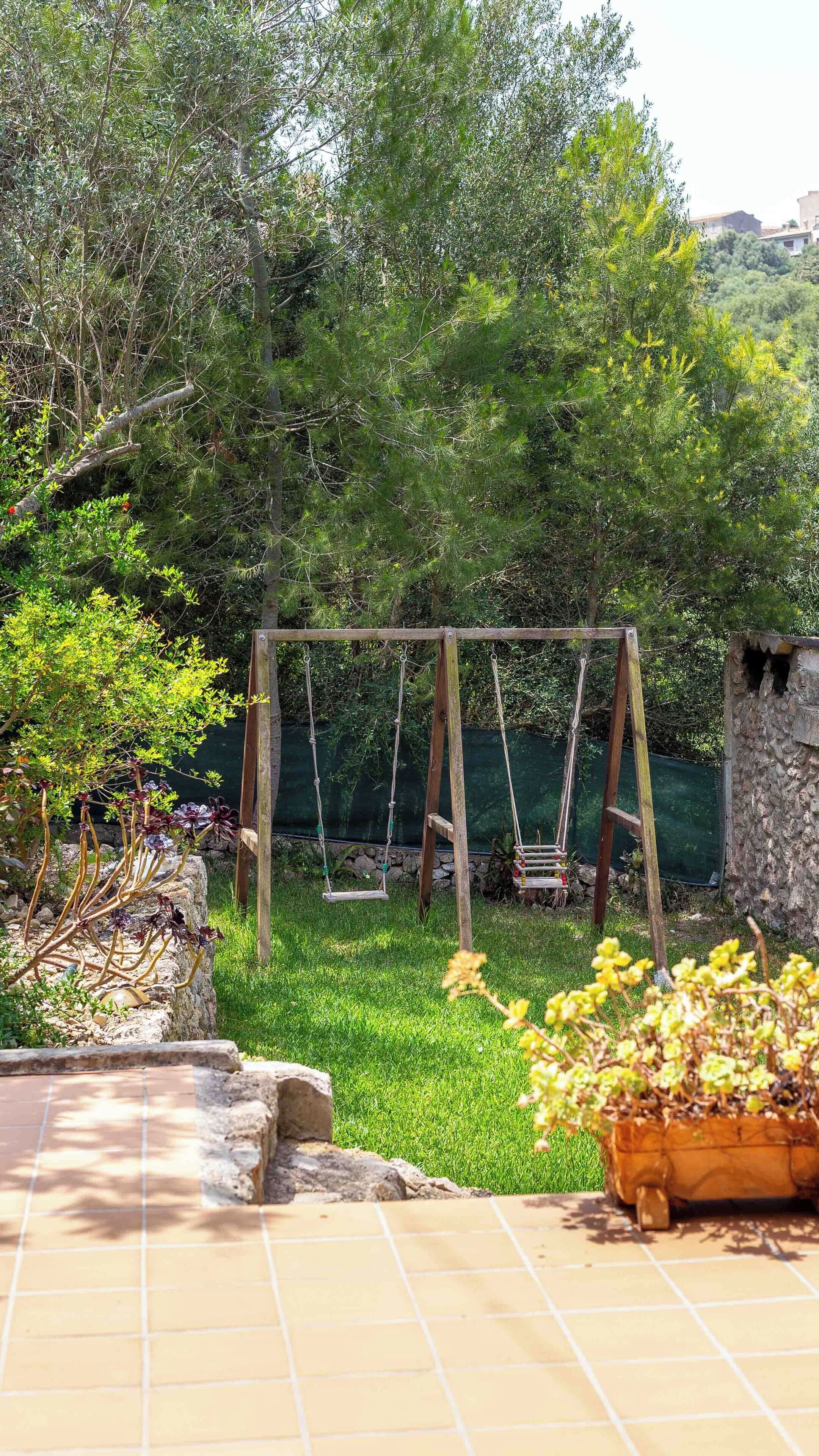 Un jardín con un conjunto de columpios de madera, un muro de piedra y una zona de patio con baldosas.