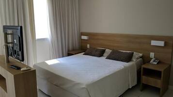 Quarto de hotel com uma cama grande, cabeceira de madeira, duas mesas de cabeceira, uma televisão e uma janela com cortinas.