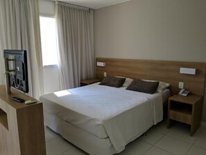 A hotel room with a large bed, wooden headboard, two bedside tables, a TV, and a window with curtains. - Apart Hotel Verano Stay Barra da Tijuca (Rio de Janeiro)
