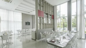 A modern dining area with white tables and chairs, a large window with a view of greenery, and a shelving unit with decorative items.