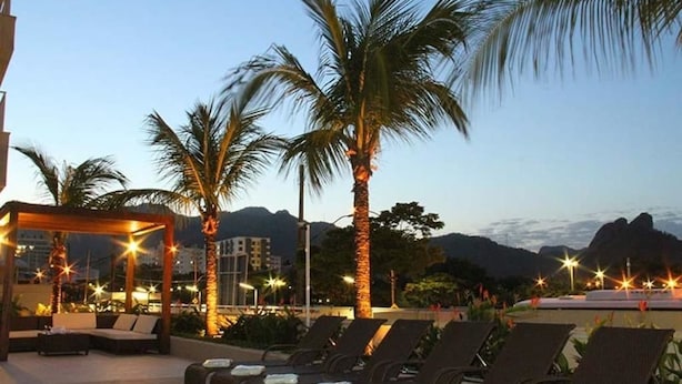 A rooftop lounge area with lounge chairs and a covered seating area, surrounded by palm trees and mountains in the background.