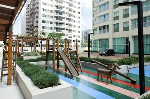 Play area with a wooden slide and climbing structures, surrounded by lush greenery and a modern apartment building with balconies and large windows.