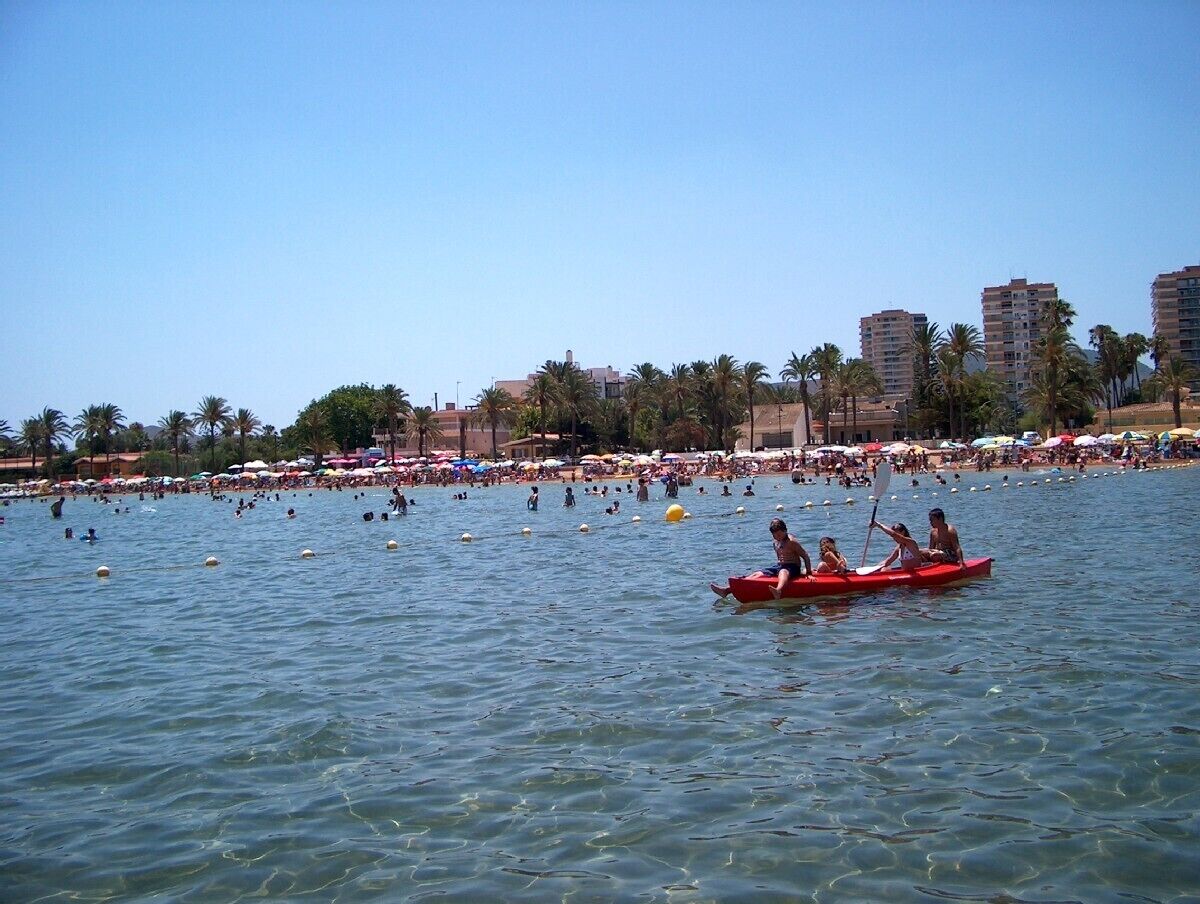 A crowded beach with people swimming and a red boat with passengers.