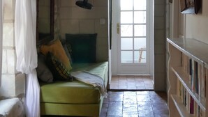 Ein schmales Zimmer mit einem Bett, einem Holzstuhl, einem Bücherregal und einer Tür zu einem anderen Raum.