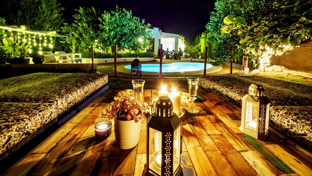 An outdoor dining area with a pool in the background, surrounded by greenery and illuminated by string lights. - Cubo's Casa Lucia Private Pool / Wifi / Air conditioning / private garden / BBQ (Alhaurín el Grande)