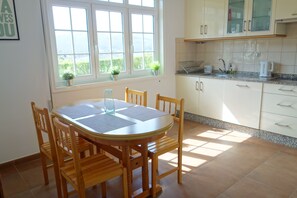 Un comedor con una mesa redonda, sillas de madera, una ventana con plantas y una cocina con armarios y encimera.