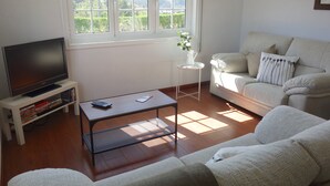 A living room with a sofa, a coffee table, a TV on a stand, and a window with a view of greenery.