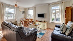 A spacious living room with leather sofas, a dining area with wooden chairs, a TV on a wooden cabinet, and large windows with curtains. - 2 Bed in Woolacombe (oc-goldbe) (Woolacombe)