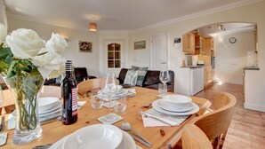 A dining area with a wooden table set for four, surrounded by chairs. There is a vase with white flowers on the table, and a bottle of wine. The room has a cozy atmosphere with a sofa and a coffee table in the background. - 2 Bed in Woolacombe (oc-goldbe) (Woolacombe)