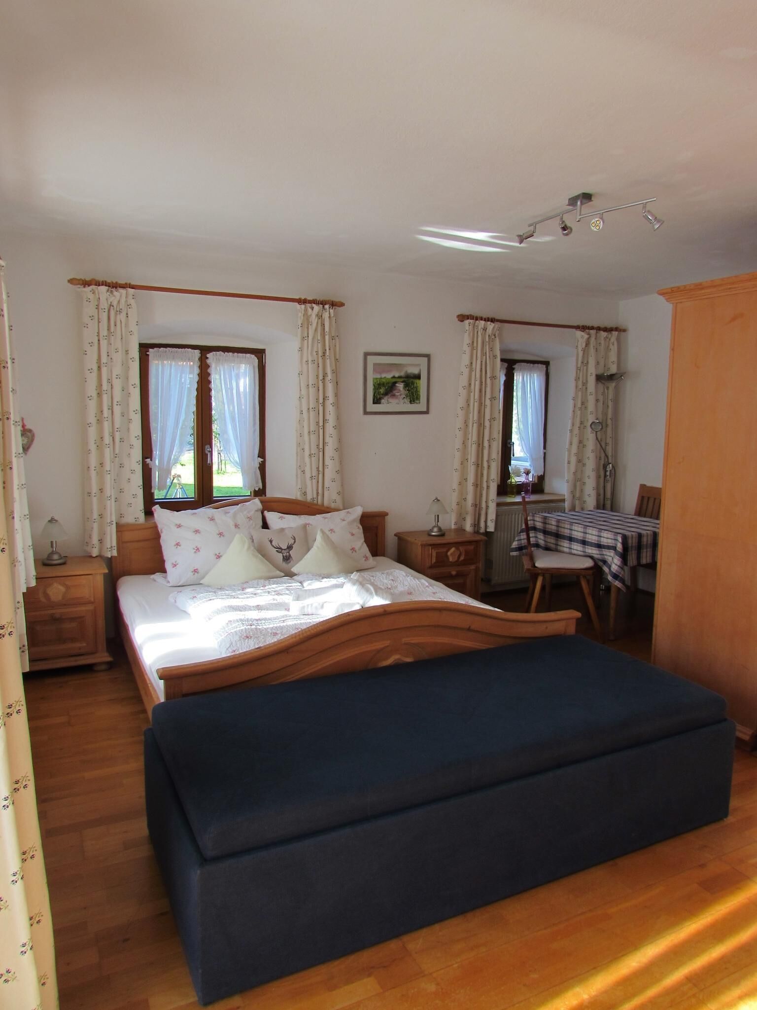 Een slaapkamer met een groot bed, een bureau en een stoel. Gordijnen hangen voor de ramen en er is een schilderij aan de muur.