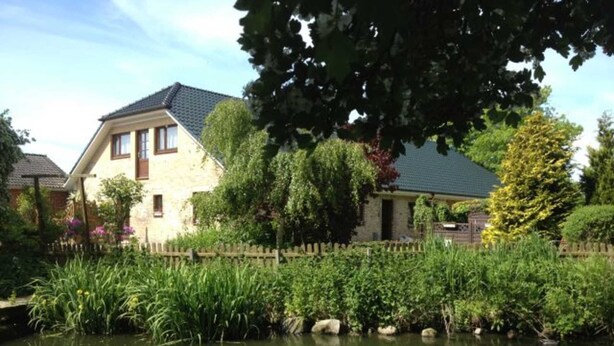 A house with a stone facade, a sloped roof, and a wooden fence surrounding a pond.
