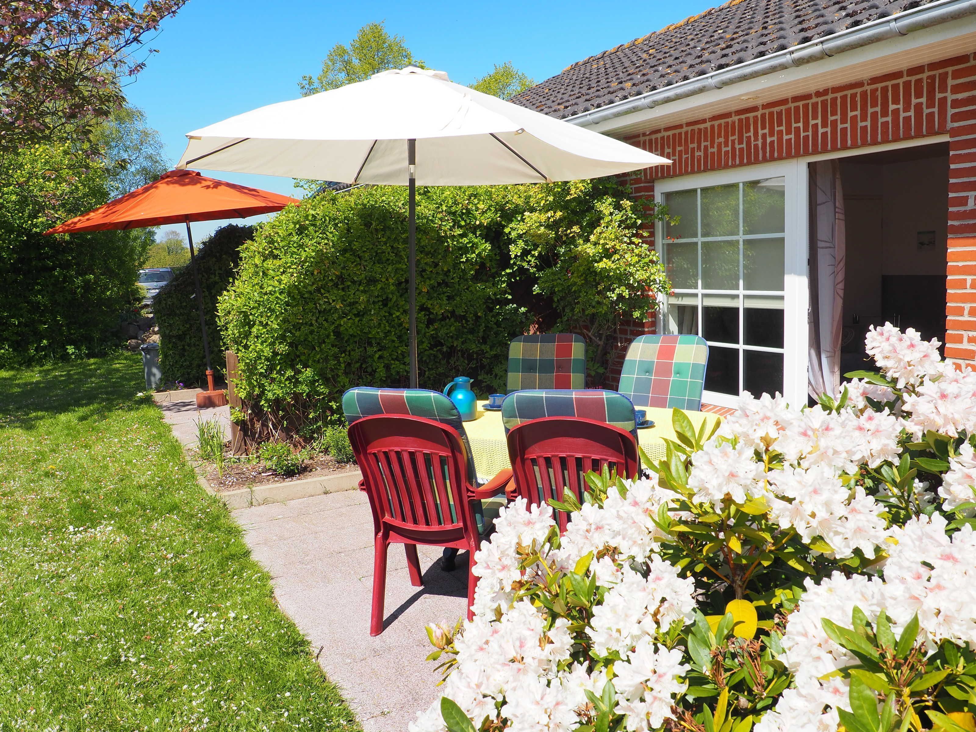 Eine Terrasse mit Gartenmöbeln, einem weißen Sonnenschirm und blühenden Blumen.