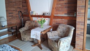 Une pièce avec un mur en briques, des poutres en bois au plafond, une table recouverte d’une nappe blanche, deux fauteuils à motifs floraux, une lampe et un vase rempli de fleurs.