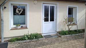 Un bâtiment avec une porte blanche, deux fenêtres, un petit porche orné de plantes et une gouttière en métal.