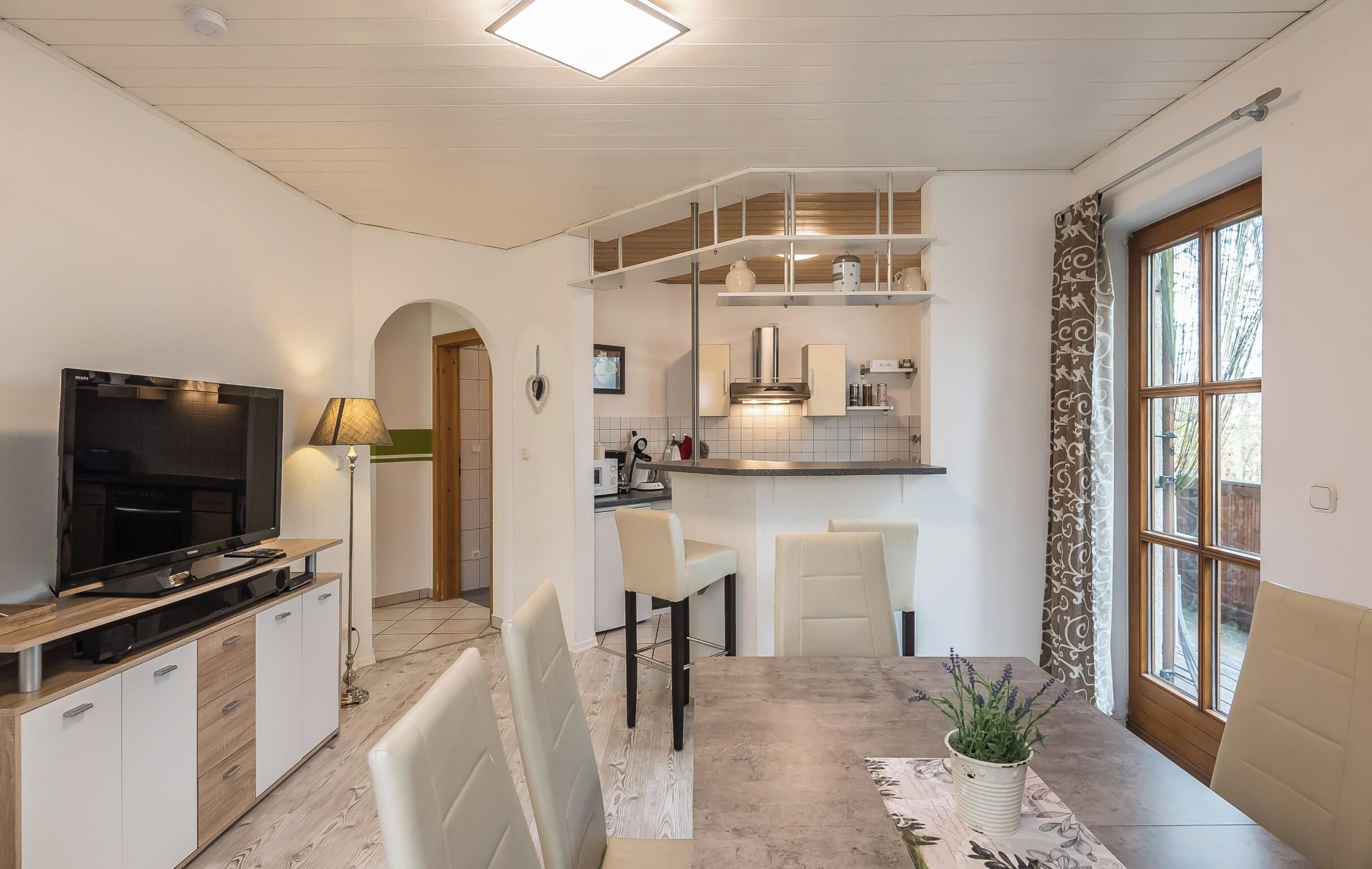 Ein modernes Wohnzimmer mit Flachbildfernseher, Essbereich mit Stühlen, Küche mit Schränken und einem großen Fenster.