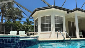 Une piscine avec un bord carrelé, un espace de repos couvert avec des chaises, et un bâtiment doté de grandes fenêtres.