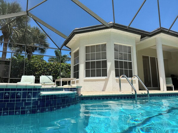 Una piscina con borde de azulejos, un área de descanso techada con sillas y un edificio con ventanas amplias.