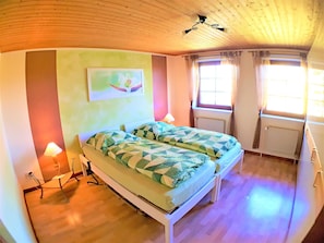Quarto com cama, duas luminárias de cabeceira, janela com cortinas, radiador e ventoinha de teto.