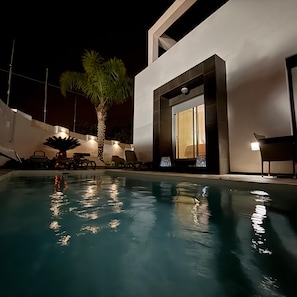 Une piscine avec des chaises longues et un palmier, entourée d’un bâtiment moderne doté de grandes fenêtres.
