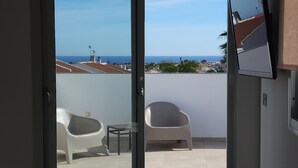 Habitación de hotel con cama, televisor montado en la pared y puerta corrediza de vidrio que da a un balcón exterior con dos sillas blancas y una mesa.