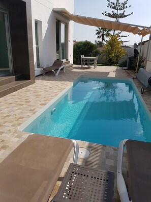 Piscina com espreguiçadeiras e uma mesa, rodeada por um terraço ladrilhado e uma área coberta com pérgola.