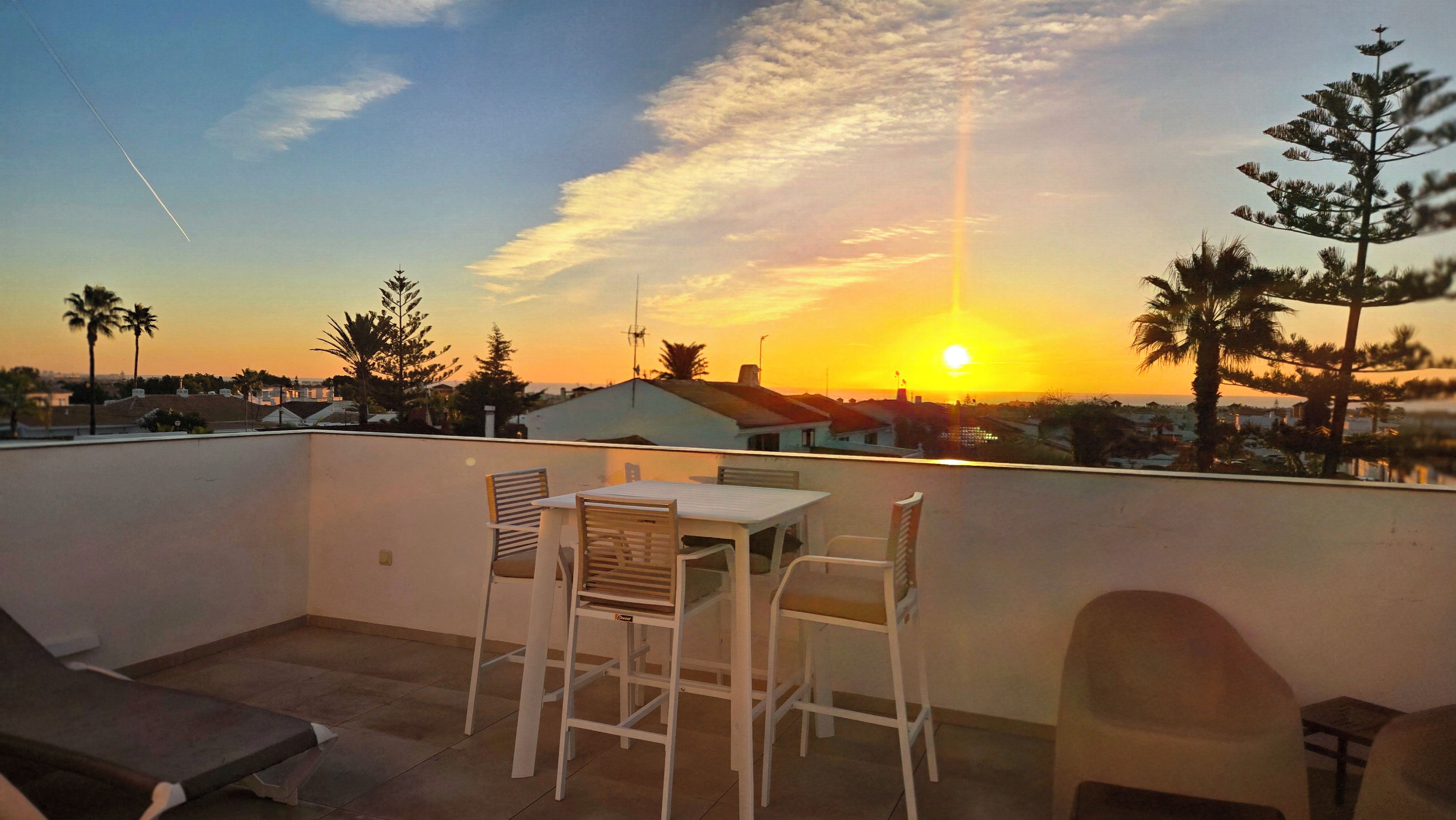 A rooftop terrace with a table and chairs, overlooking a residential area during sunset.
