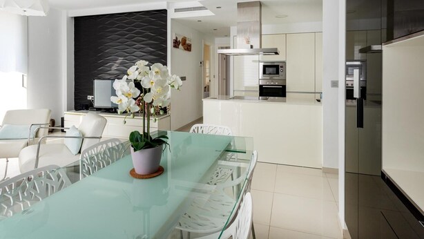 Cocina moderna con electrodomésticos de acero inoxidable, una mesa de vidrio y una orquídea en maceta, además de una silla blanca.
