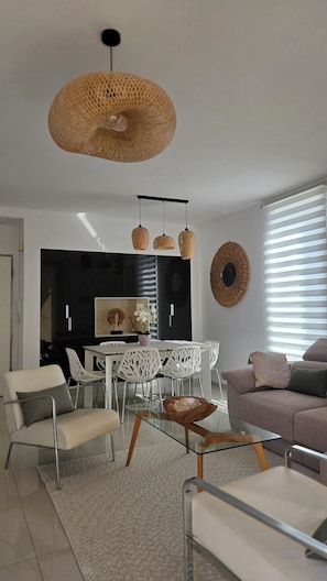 Un salon moderne avec une table basse en verre, des chaises blanches, un canapé et un coin repas comprenant un buffet noir et des chaises blanches.