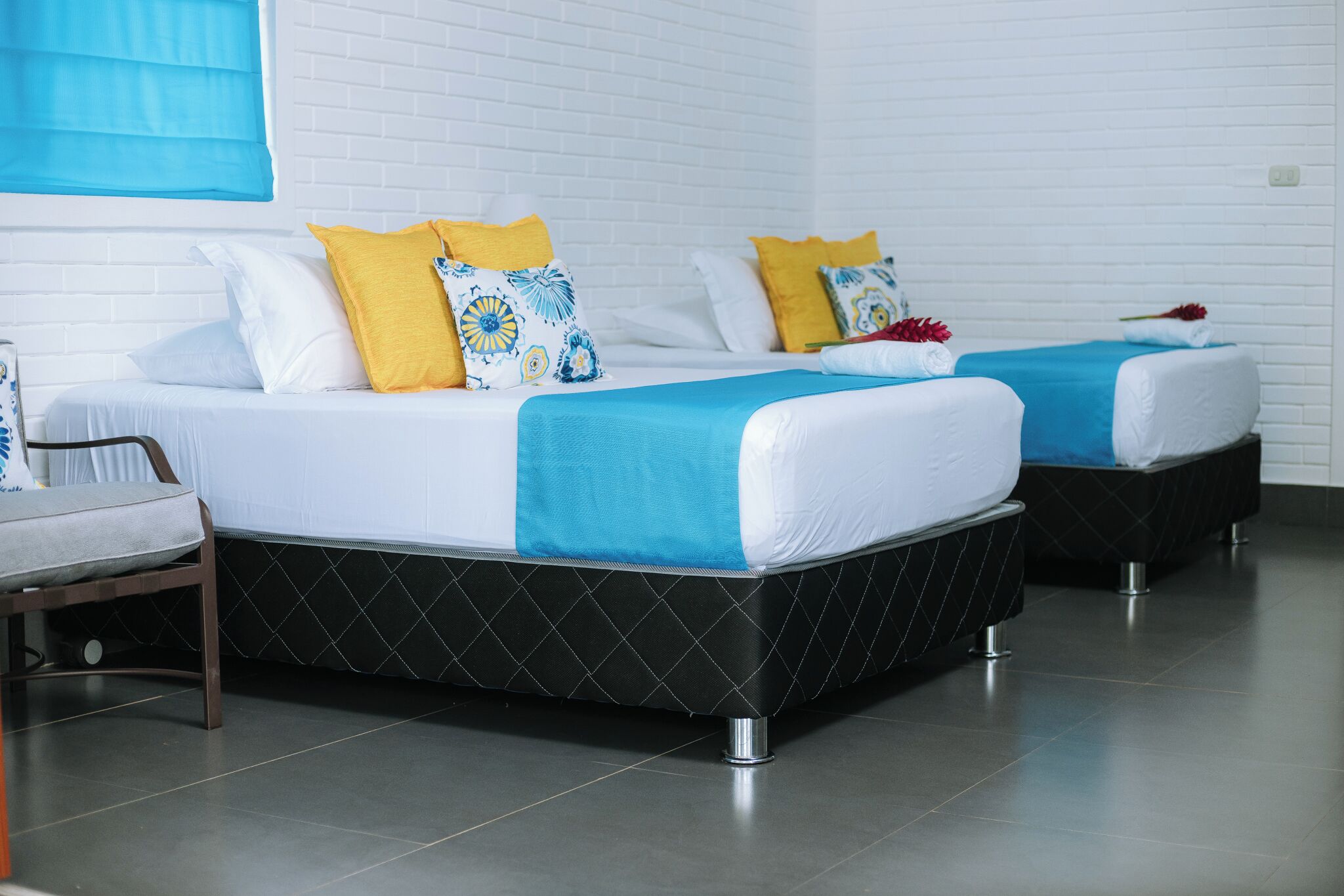 Dos camas con ropa de cama blanca y azul, una persiana azul y una pared de ladrillo blanco.