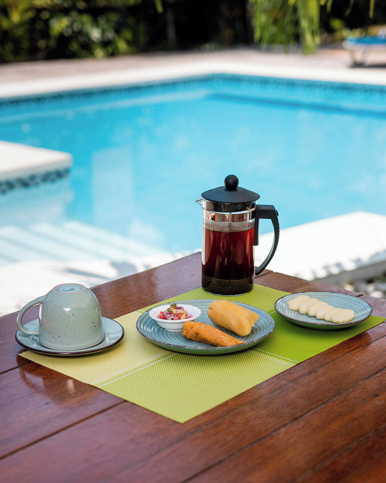 Une table avec une cafetière, une théière, des assiettes remplies de nourriture et une boisson, le tout installé au bord d’une piscine.