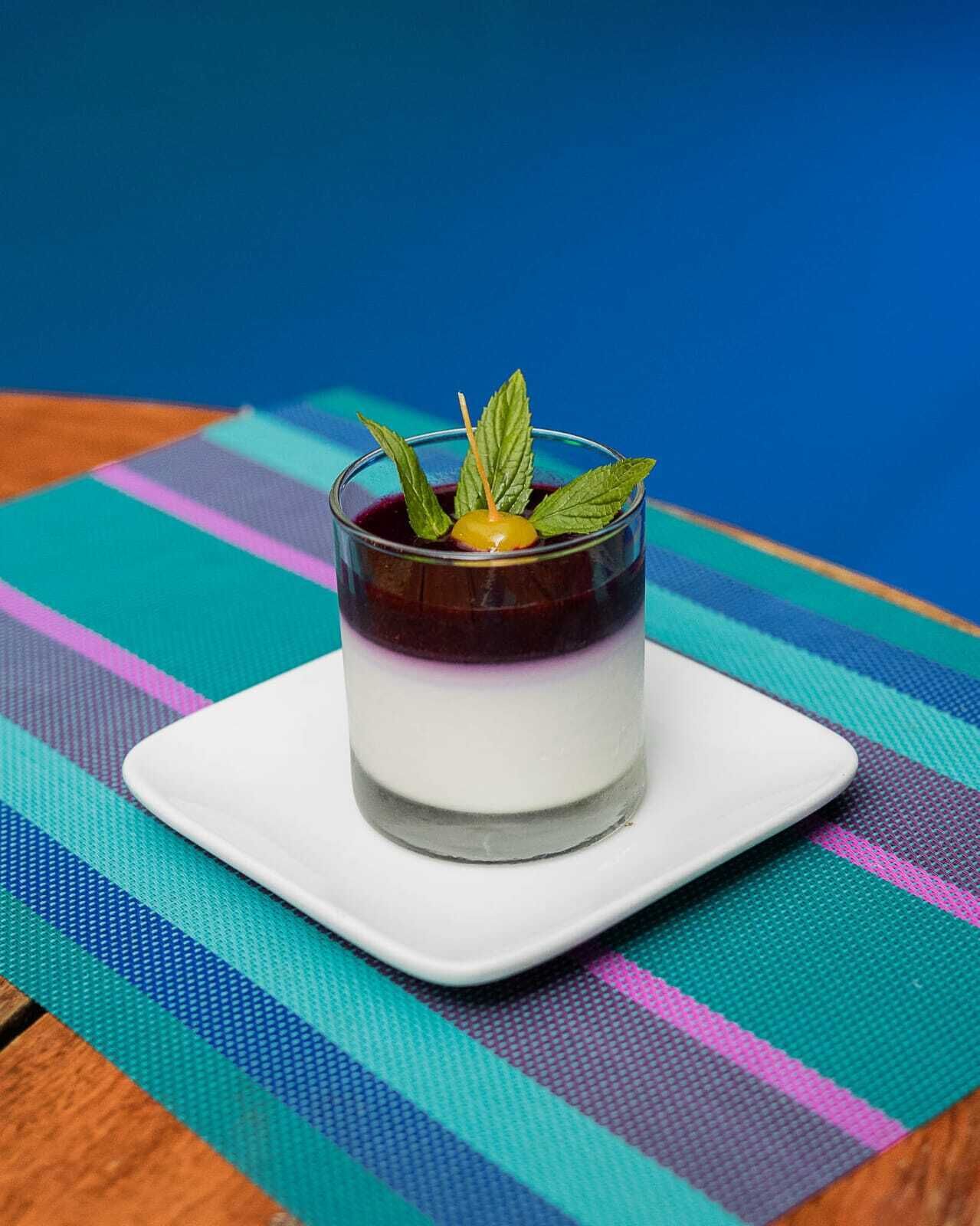 Uma sobremesa em copo sobre um prato branco, colocada num tapete colorido listrado.