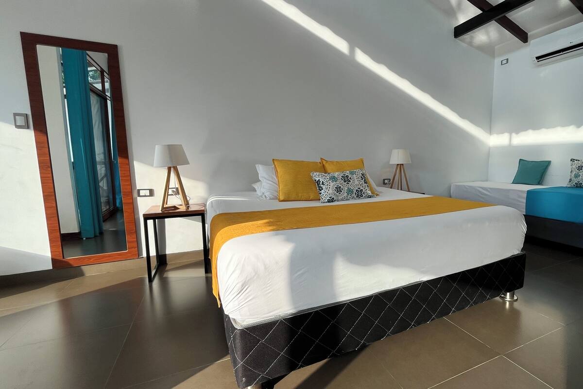 A modern hotel room with a large bed, a smaller sofa, a nightstand with a lamp, and a large mirror.