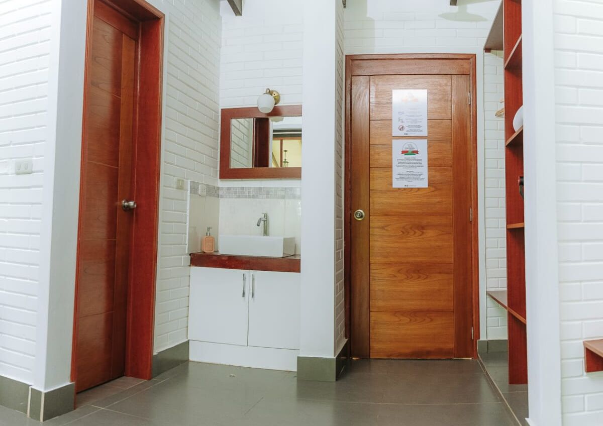 Un baño con puerta de madera, un lavamanos blanco con espejo arriba y pared cubierta de azulejos blancos.